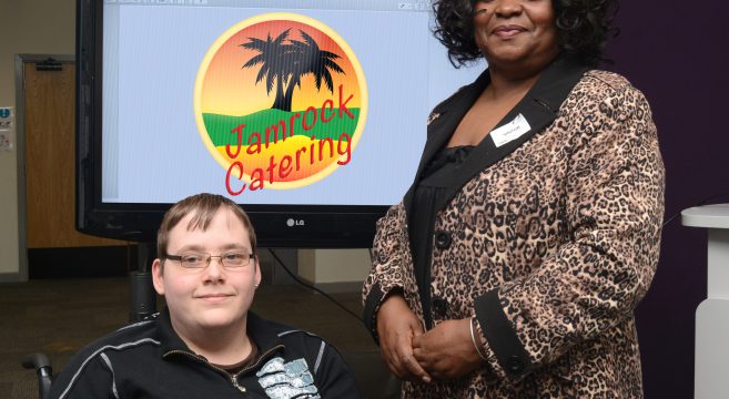 Male student in wheelchair with woman standing in front of Jamrock Catering logo on screen