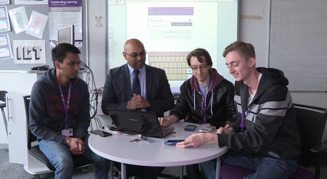 Three students and lecturer sitting around table looking at phones and laptop