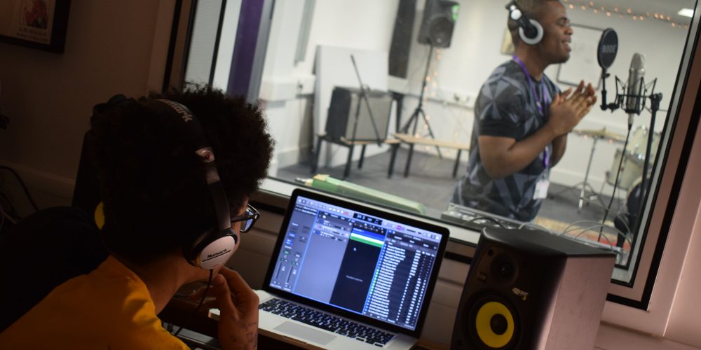Student singing in recording studio whilst another student listens in on his headphones